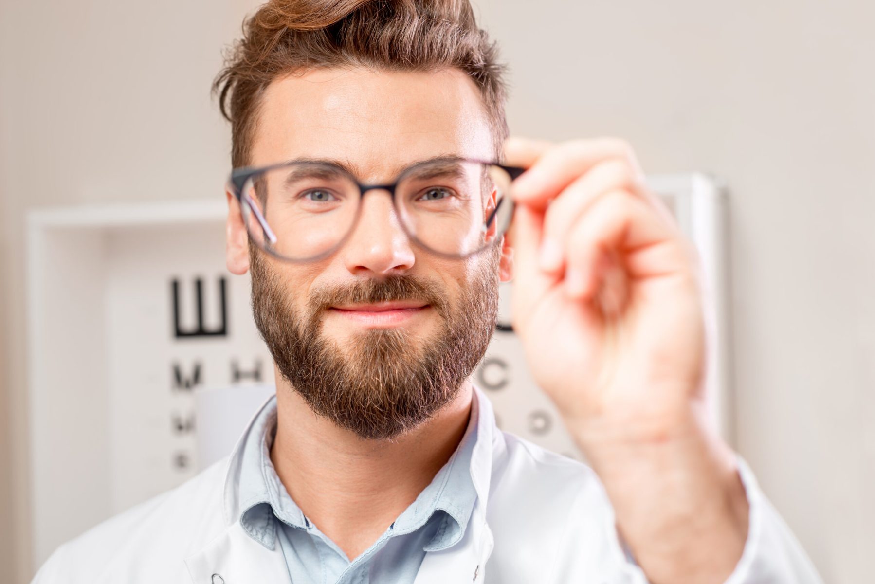 Handsome ophthalmologist looking through the glasses on the eye chart background in the cabinet